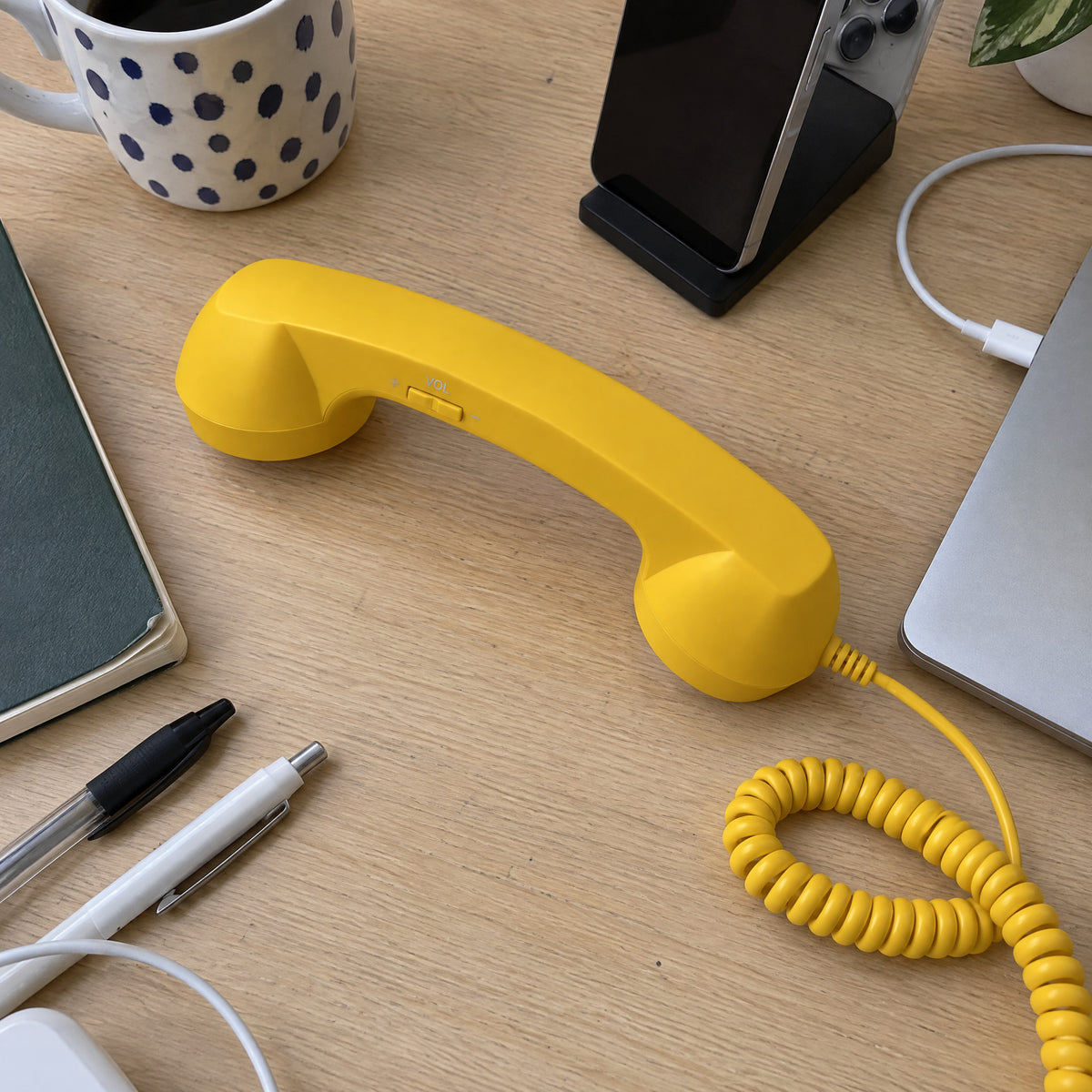 Yellow retro handset with coiled cord on a wooden desk beside a coffee mug, notebook, pens, phone stand and laptop