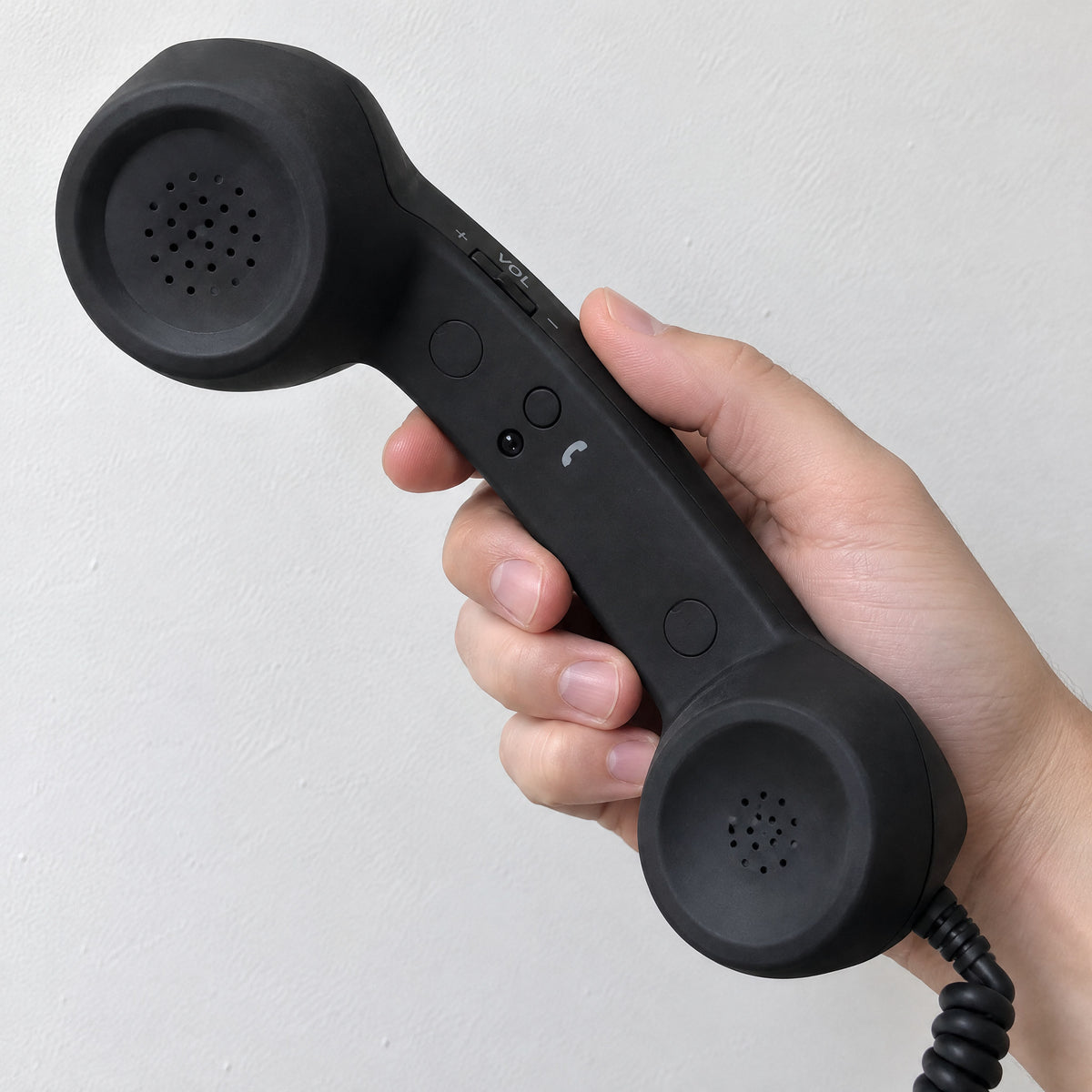 Hand holding a black retro handset, showing the + VOL - volume rocker, buttons and attached black coiled cord