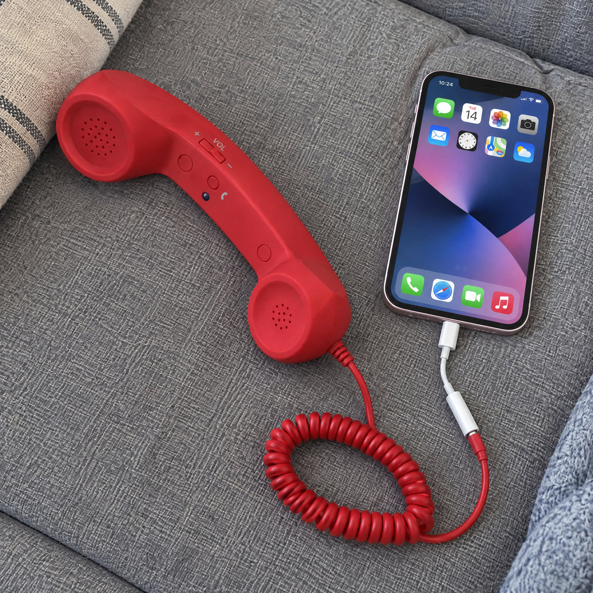 Red retro handset with coiled cord plugged into an iPhone through a white Lightning adapter, resting on a grey sofa