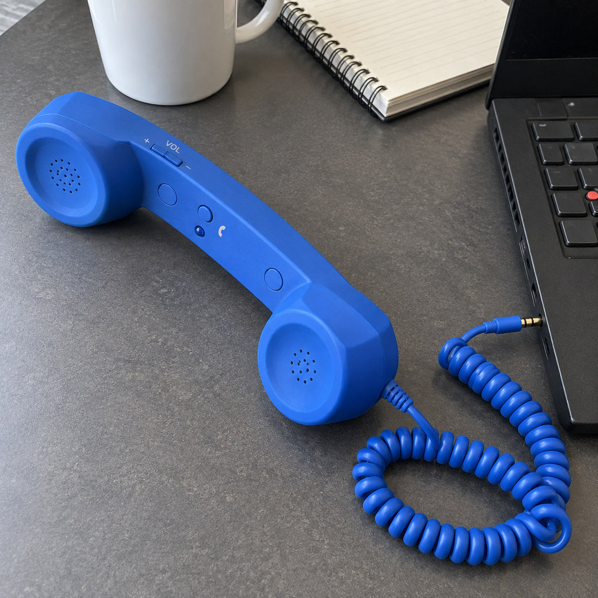 Blue retro handset with coiled cord plugged by 3.5 mm jack into a black laptop on a grey desk, with mug and notebook